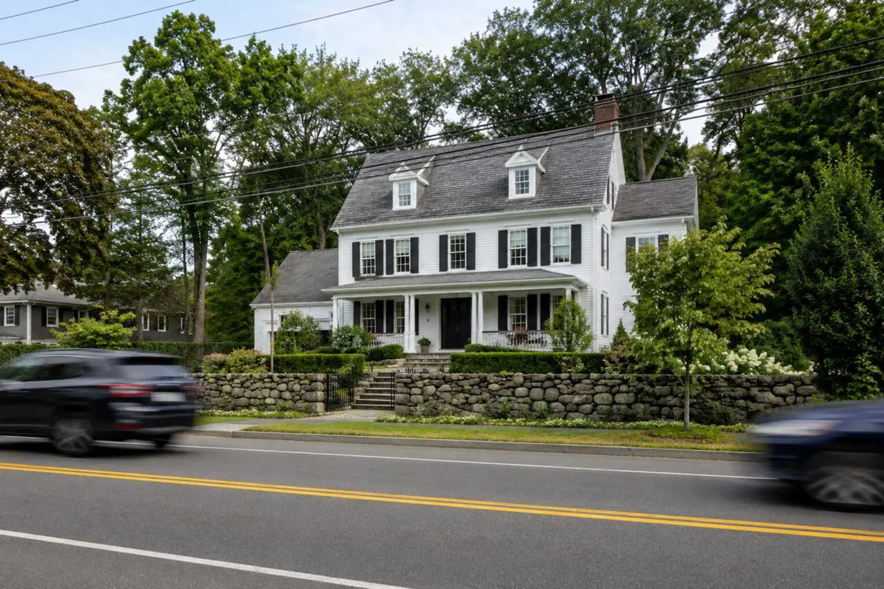 A New England-style home on a busy road shows how stone walls, layered planting, and a defined entry can soften exposure while preserving curb appeal.