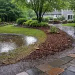 Standing water collects in a lawn depression while a light runoff line follows the edge of a mulched bed and stone wall, illustrating a common residential drainage issue on a New England property.