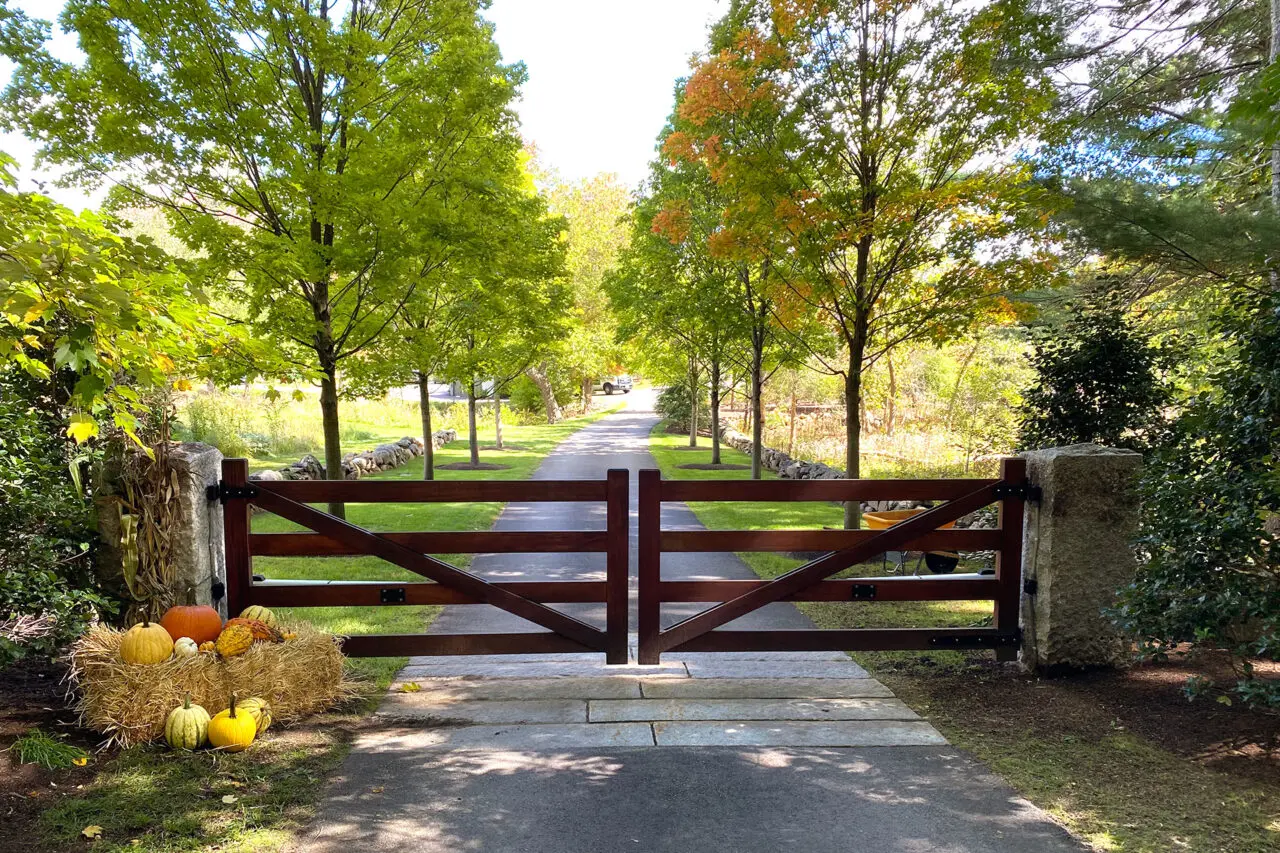 A handcrafted wood gate frames a tree-lined drive, creating a formal yet rural arrival sequence that sets the tone for this historic New England estate.