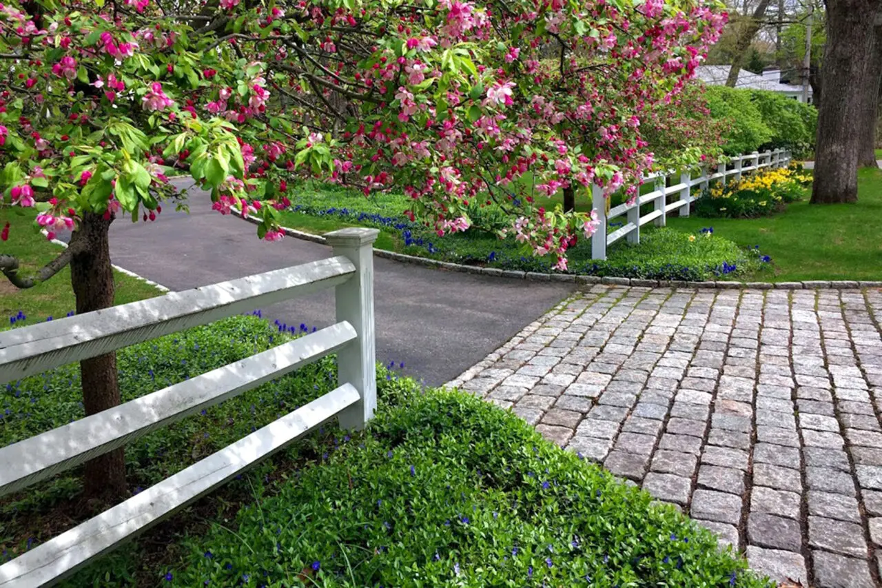 A cobblestone driveway framed by a flowering tree and white fence delivers a charming spring entry, combining texture, durability, and traditional detailing.