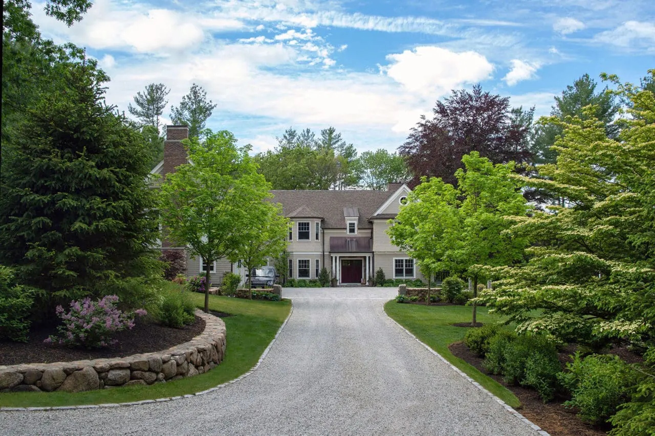A straight gravel driveway is reinforced by parallel stone walls and a tree-lined approach, delivering clear sightlines, strong geometry, and a timeless sense of order.