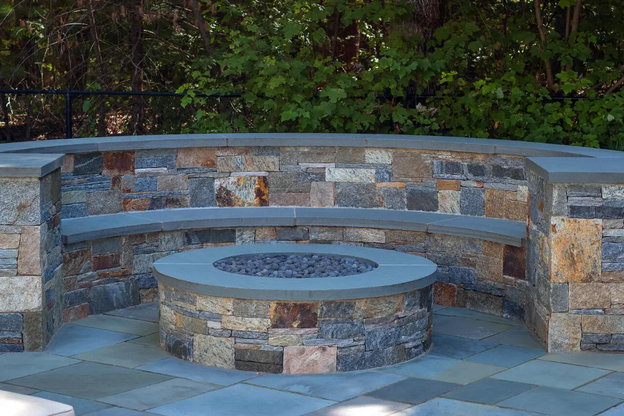A round gas fire pit with a curved stone seating wall offers consistent heat and low maintenance, creating a comfortable gathering space that feels both elegant and highly functional.