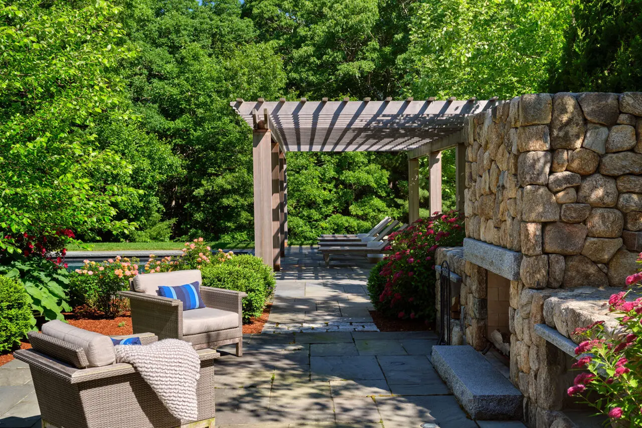 A stone outdoor fireplace beneath a pergola defines this lounge patio, adding shade, structure, and warmth while creating a year-round destination for relaxed seating and conversation.