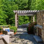 A stone outdoor fireplace beneath a pergola defines this lounge patio, adding shade, structure, and warmth while creating a year-round destination for relaxed seating and conversation.