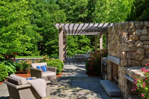 A stone outdoor fireplace beneath a pergola defines this lounge patio, adding shade, structure, and warmth while creating a year-round destination for relaxed seating and conversation.