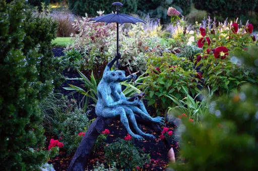 A bronze frog statue holding an umbrella brings humor and personality to the planting bed, its rich patina contrasting beautifully with fresh greens and flowers.