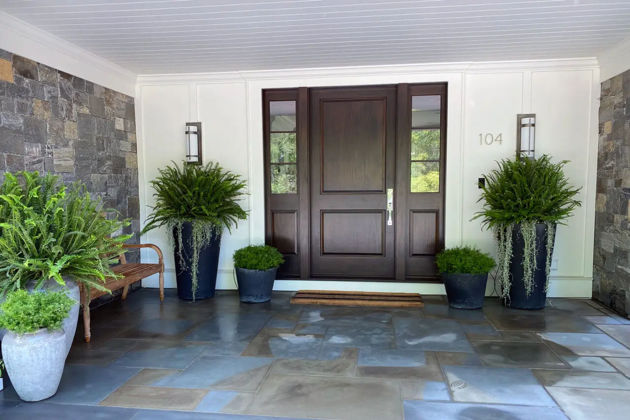 Multiple planters are grouped along a covered entry, layering ferns and trailing vines to create rhythm, depth, and a cohesive summer display that softens architecture with low maintenance care.