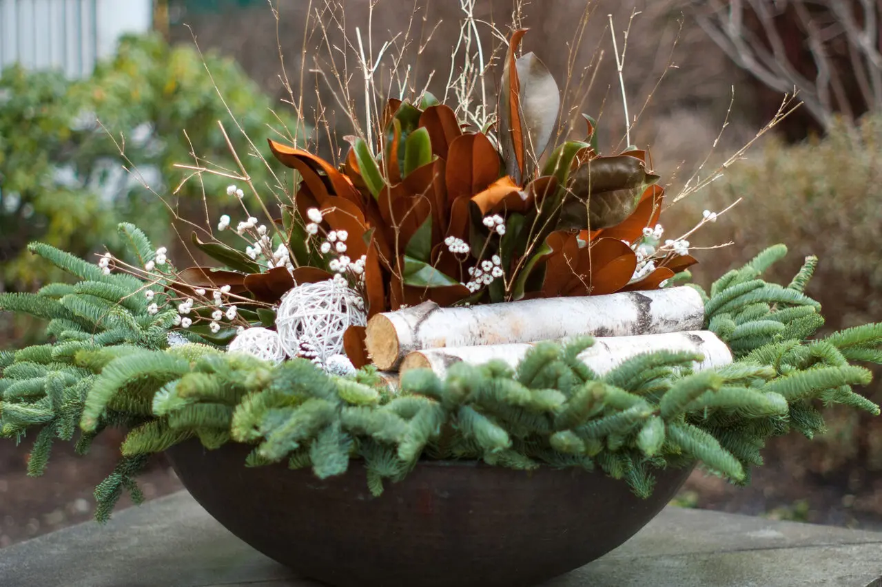 A shallow metal bowl holds a winter arrangement of evergreen cuttings and birch logs, functioning as a compact seasonal centerpiece.