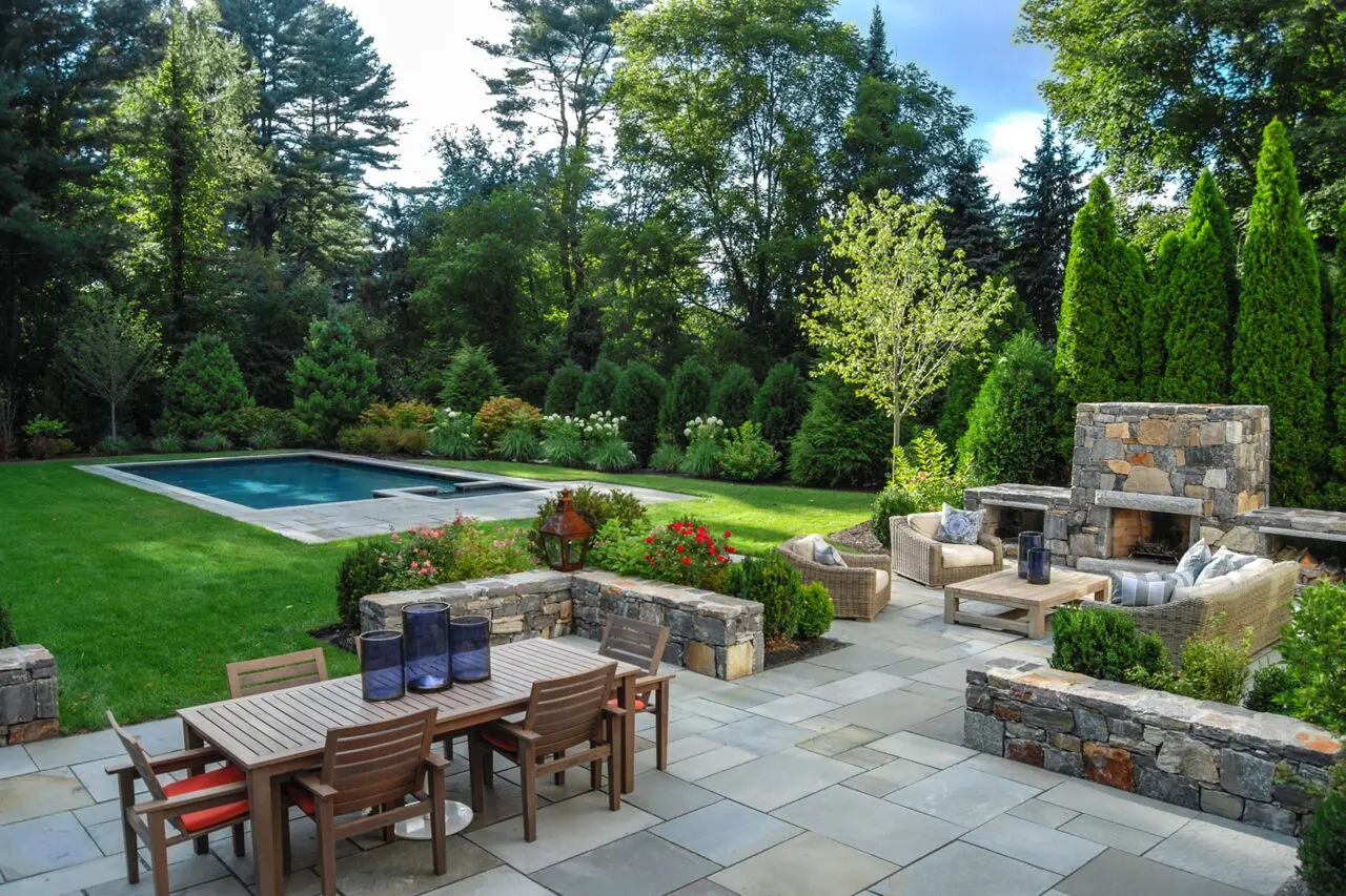 A rectangular stone pool anchored by a bluestone terrace and outdoor fireplace, creating a refined entertaining space that blends structured hardscape with soft planting for year-round appeal in Weston.