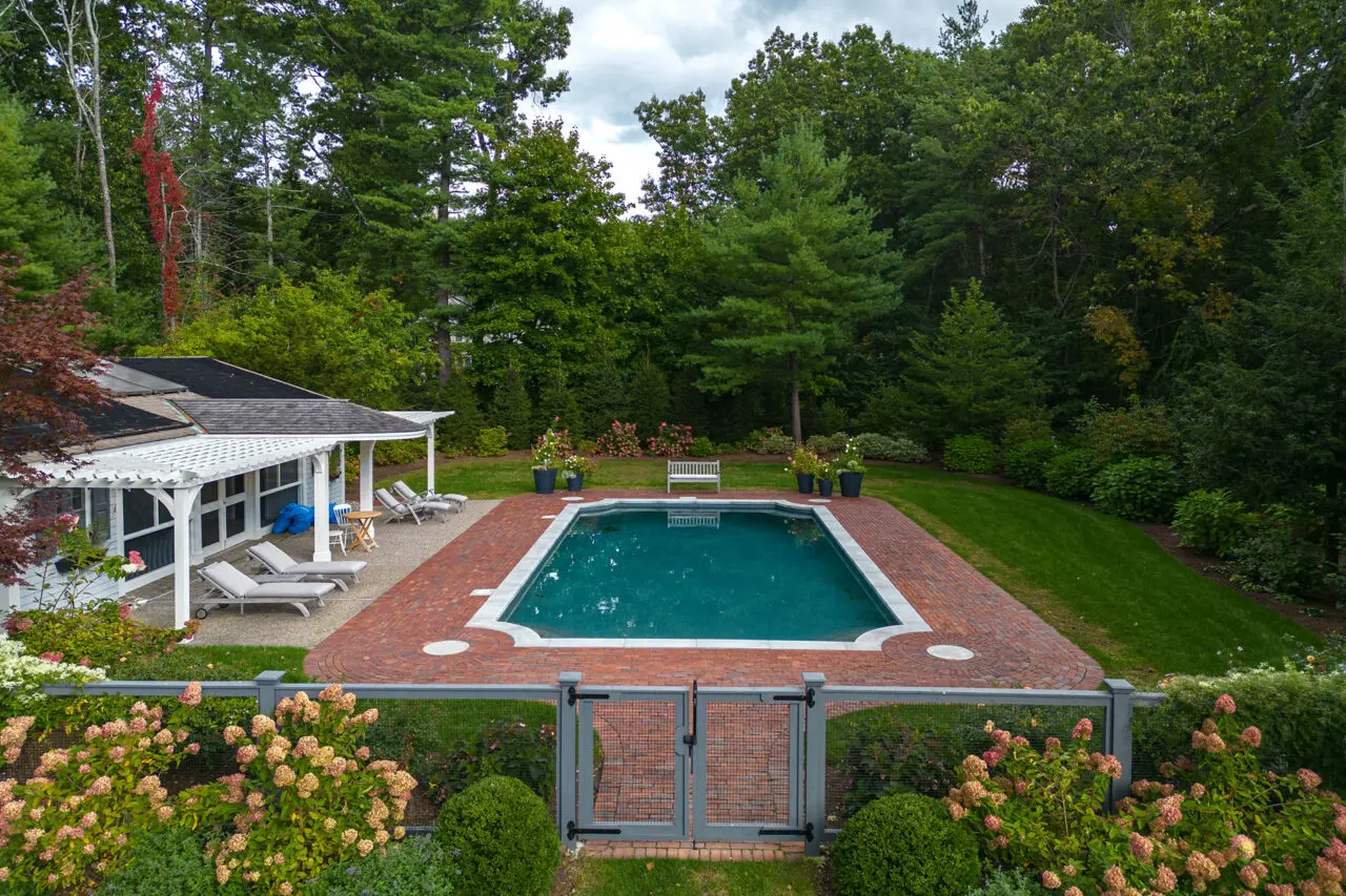 A classic rectangular pool set within a brick patio and woodland backdrop, offering a timeless New England look that balances clean geometry with natural privacy and garden integration.