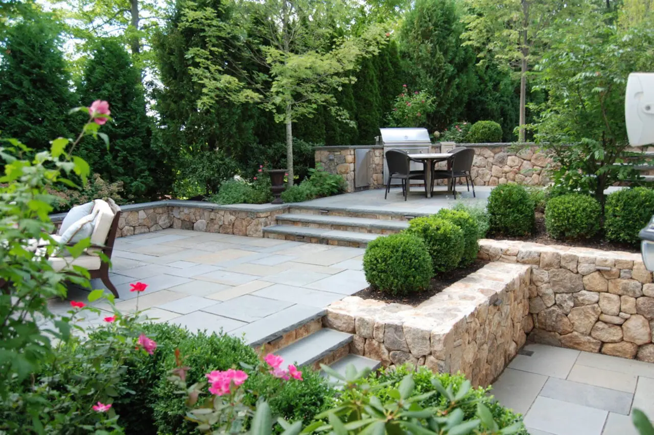 Stone retaining walls surround the bluestone patio and outdoor kitchen, with shrubs and hedges providing privacy, wind protection, and a lush backdrop.
