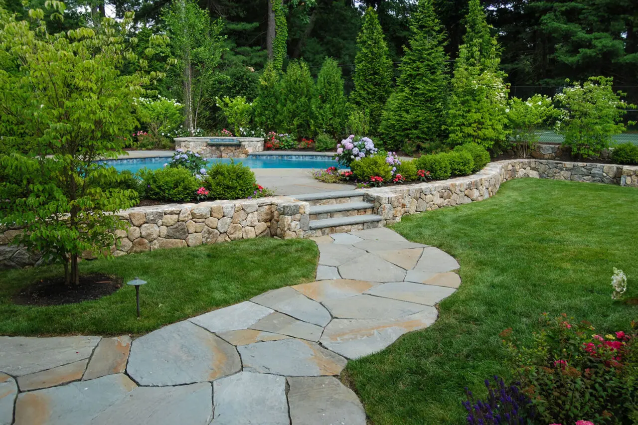 A curved flagstone walkway meets a stone retaining wall near the pool terrace, with shrubs and hedges softening curves and enhancing flow.