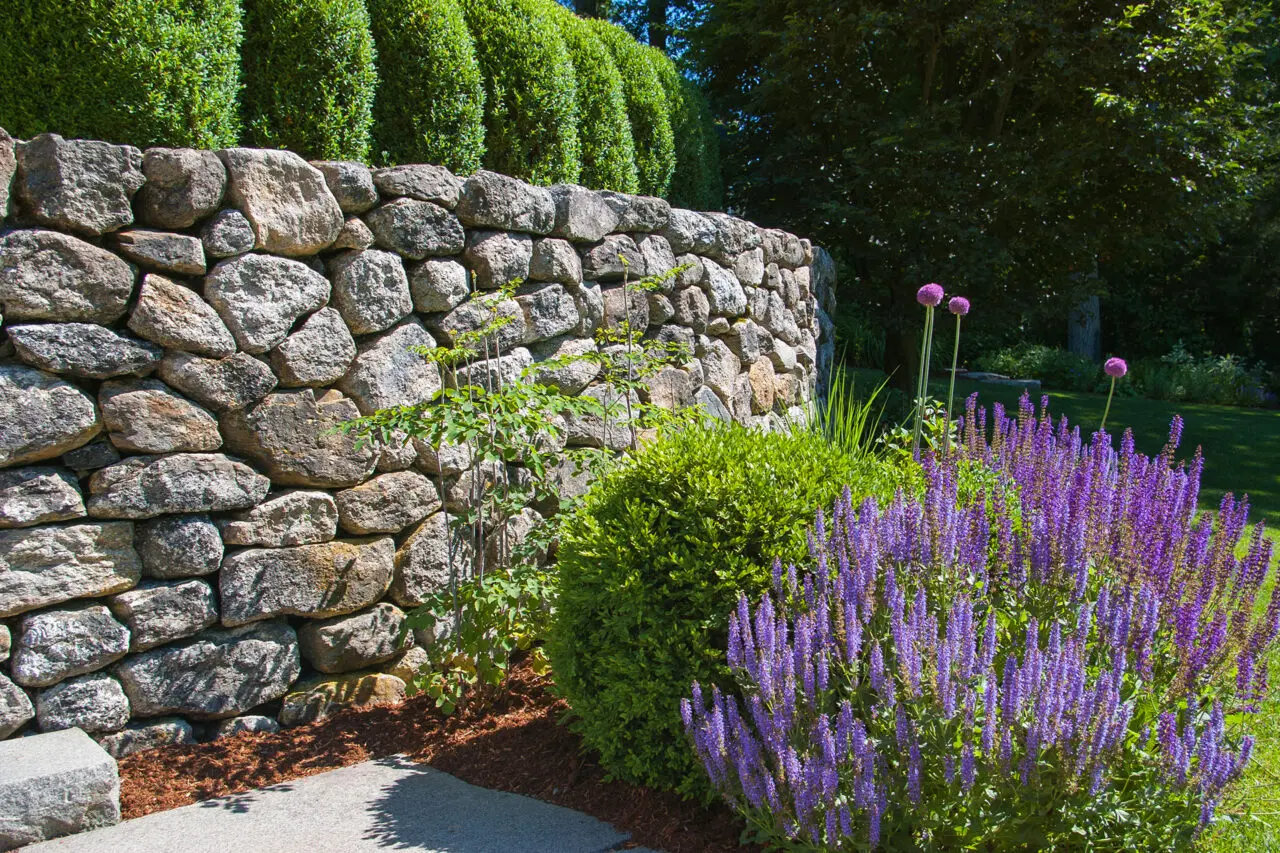 A dry-laid fieldstone retaining wall defines the garden edge, using irregular, hand-set stone to manage a subtle grade change while creating a low, natural boundary with a relaxed, timeless character.