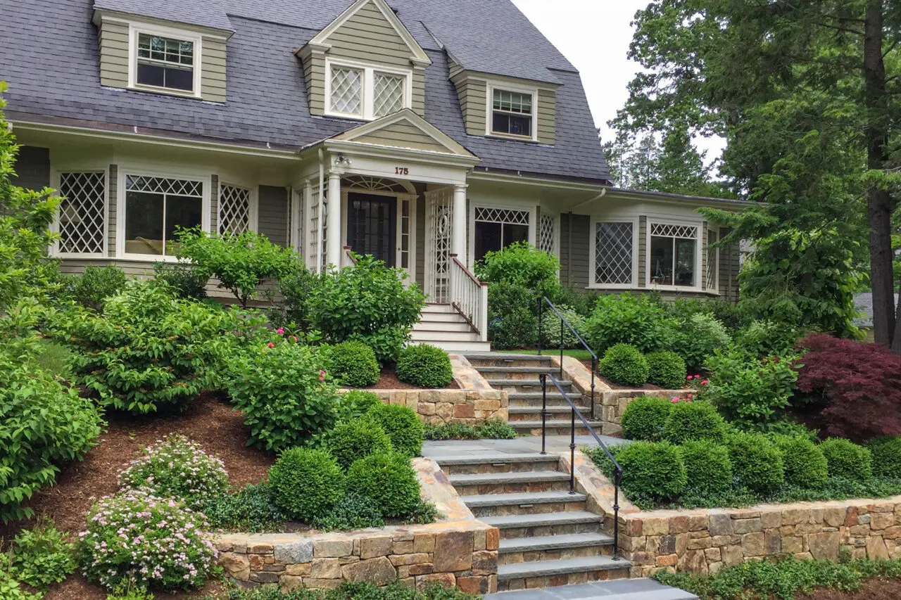Fieldstone retaining walls paired with granite steps guide movement along a hillside path, using shrubs and hedges to stabilize slopes and frame the route.