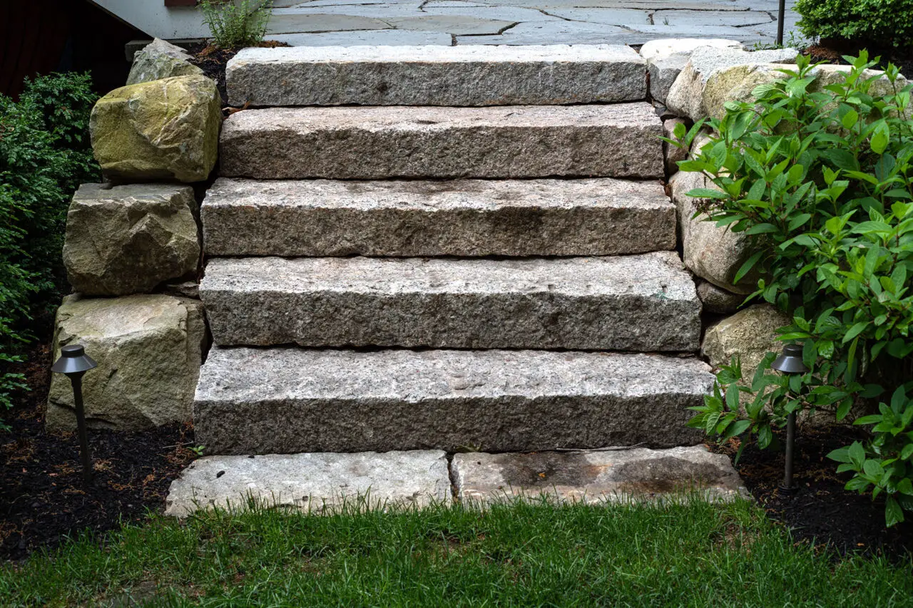 Granite block steps with boulder edging highlight changes in elevation, while nearby shrubs and hedges help frame lighting details and soften the hardscape.