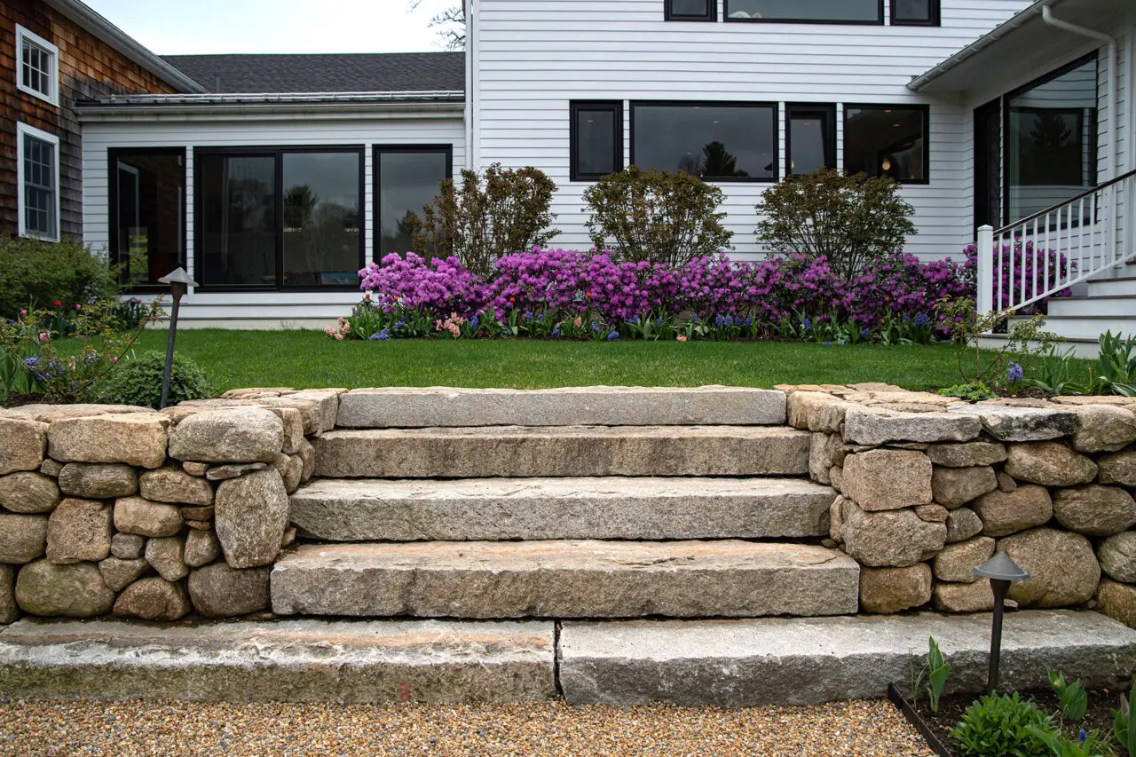 These granite steps are set into fieldstone walls near the foundation, where carefully scaled shrubs and hedges help transition between architecture and landscape.
