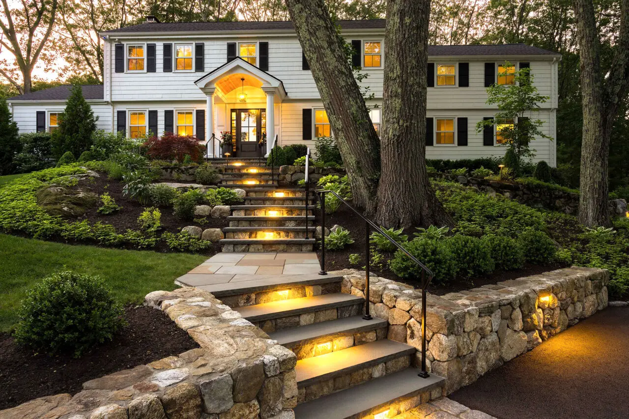 Illuminated stone steps climb a hillside approach, combining safety and visual appeal while adjacent shrubs and hedges frame the path and enhance nighttime presence.