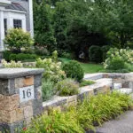 A low stone entry wall paired with granite steps establishes a clear arrival sequence, with adjacent hedges and foundation shrubs adding definition and a welcoming sense of enclosure.
