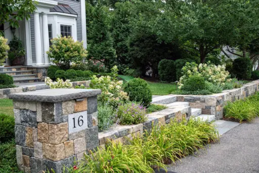 A low stone entry wall paired with granite steps establishes a clear arrival sequence, with adjacent hedges and foundation shrubs adding definition and a welcoming sense of enclosure.