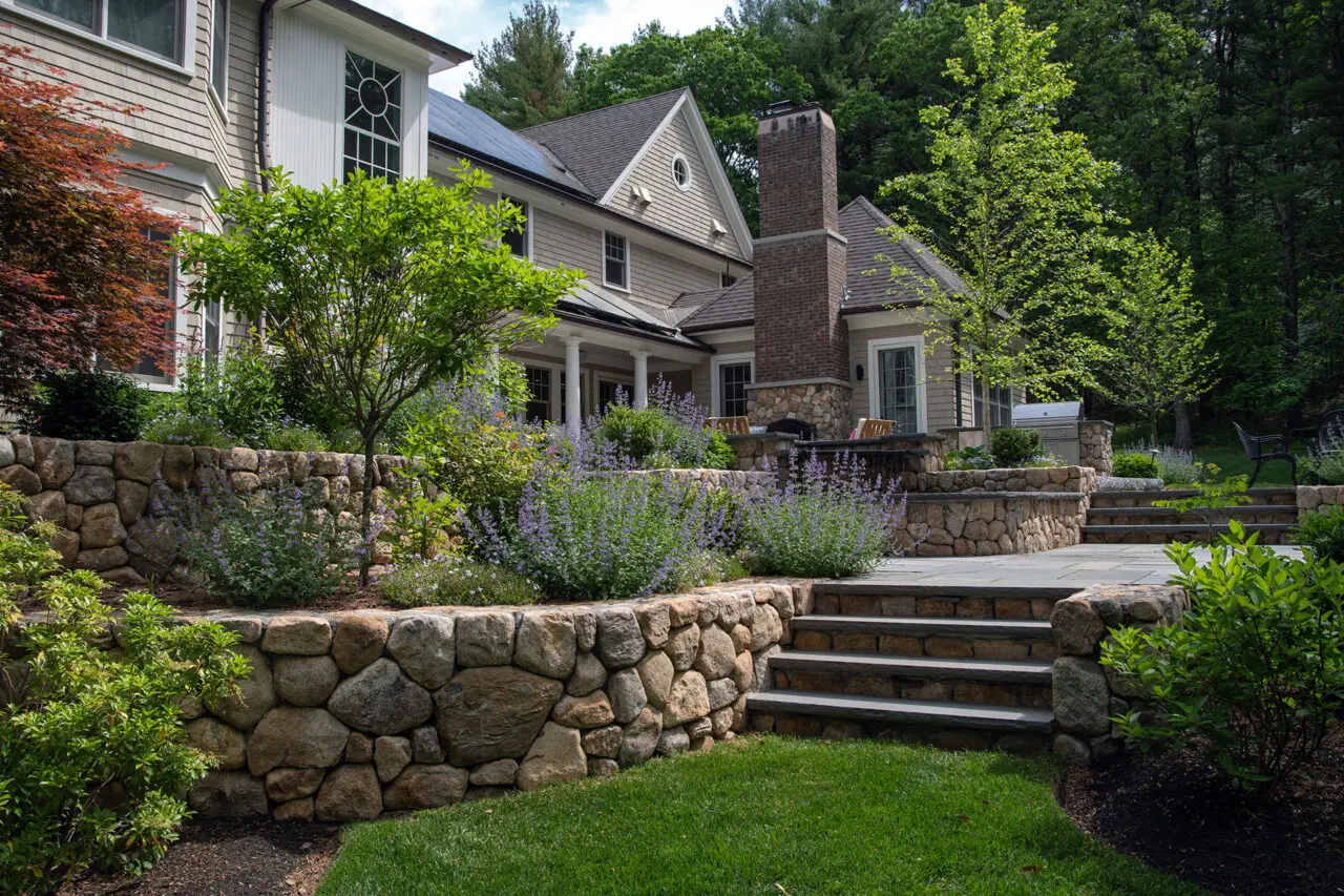 Tiered fieldstone walls paired with bluestone steps create a strong visual connection between the patio and surrounding planting, using layered stonework to manage grade while framing shrubs and low hedges that soften transitions between outdoor rooms.