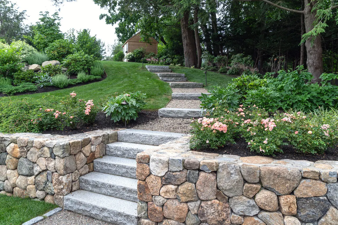 Curved stone garden walls trace the lawn edge, guiding movement through the landscape and creating planting pockets for shrubs and hedges that soften the geometry and add seasonal interest.