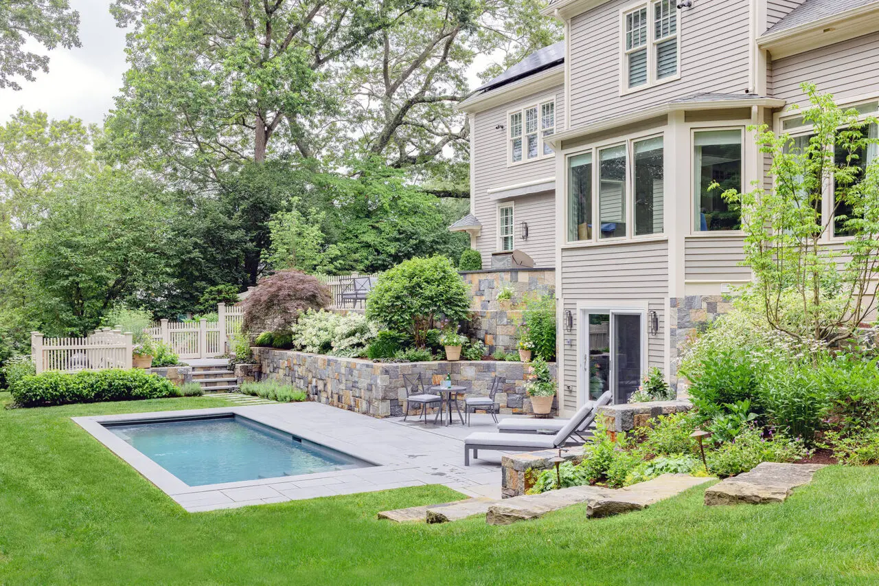 Stacked stone retaining walls step down toward the pool terrace, offering both erosion control and visual rhythm while allowing shrubs and evergreen hedging to be integrated for privacy and scale.