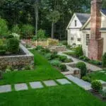 A traditional fieldstone wall anchors the front yard, its weathered texture and warm tones complemented by neatly shaped shrubs and hedges that reinforce symmetry and provide year-round structure near the home.