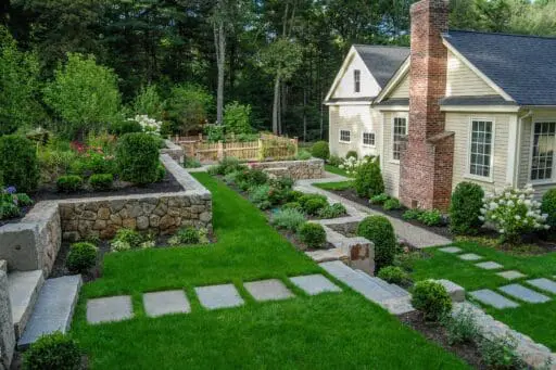 A traditional fieldstone wall anchors the front yard, its weathered texture and warm tones complemented by neatly shaped shrubs and hedges that reinforce symmetry and provide year-round structure near the home.