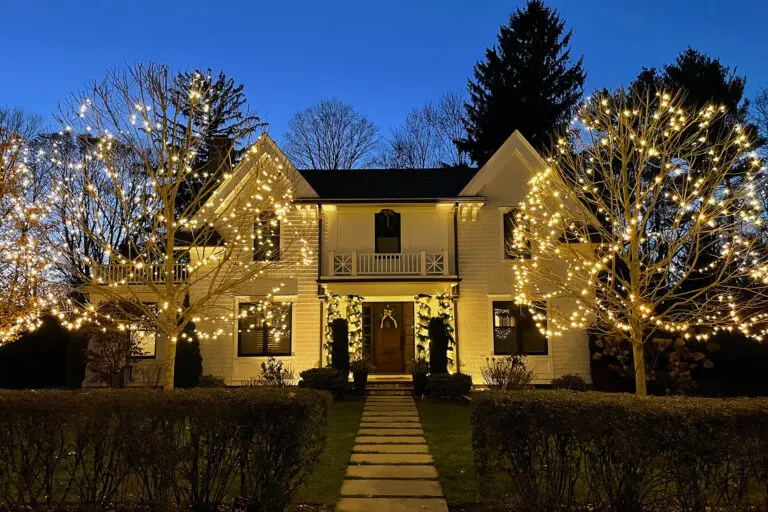 Holiday lighting highlights the front yard and entry of a New England home, creating a warm, welcoming evening presence.