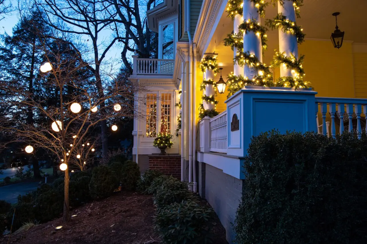A cozy winter evening scene: a classic wrap-around porch warmly illuminated with soft string lights intertwined around columns and lush evergreen garlands, casting a welcoming glow against the twilight sky and highlighting the architectural charm of the home.