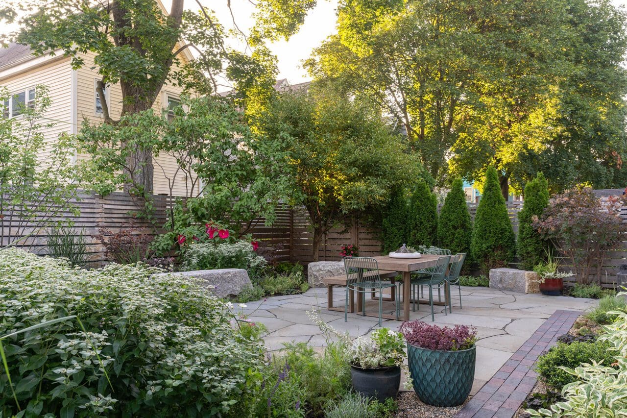 An outdoor dining area arranged like an interior room, with defined seating, structured planting, and durable stone paving that creates a comfortable, purposeful extension of the home.