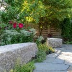 A curved bluestone garden path bordered by native plantings, flowering shrubs, and mature trees, with large granite seating stones and a modern wooden privacy fence creating a quiet, shaded backyard retreat.
