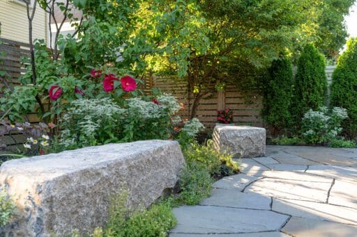 A curved bluestone garden path bordered by native plantings, flowering shrubs, and mature trees, with large granite seating stones and a modern wooden privacy fence creating a quiet, shaded backyard retreat.