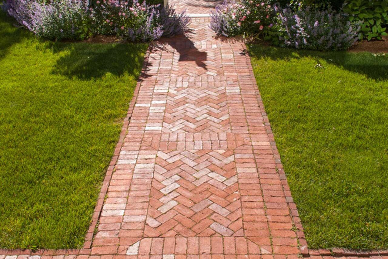 Cambridge, MA - A classic herringbone brick walkway creates a welcoming approach to a historic garden gate, framed by lush perennial plantings and lawn.