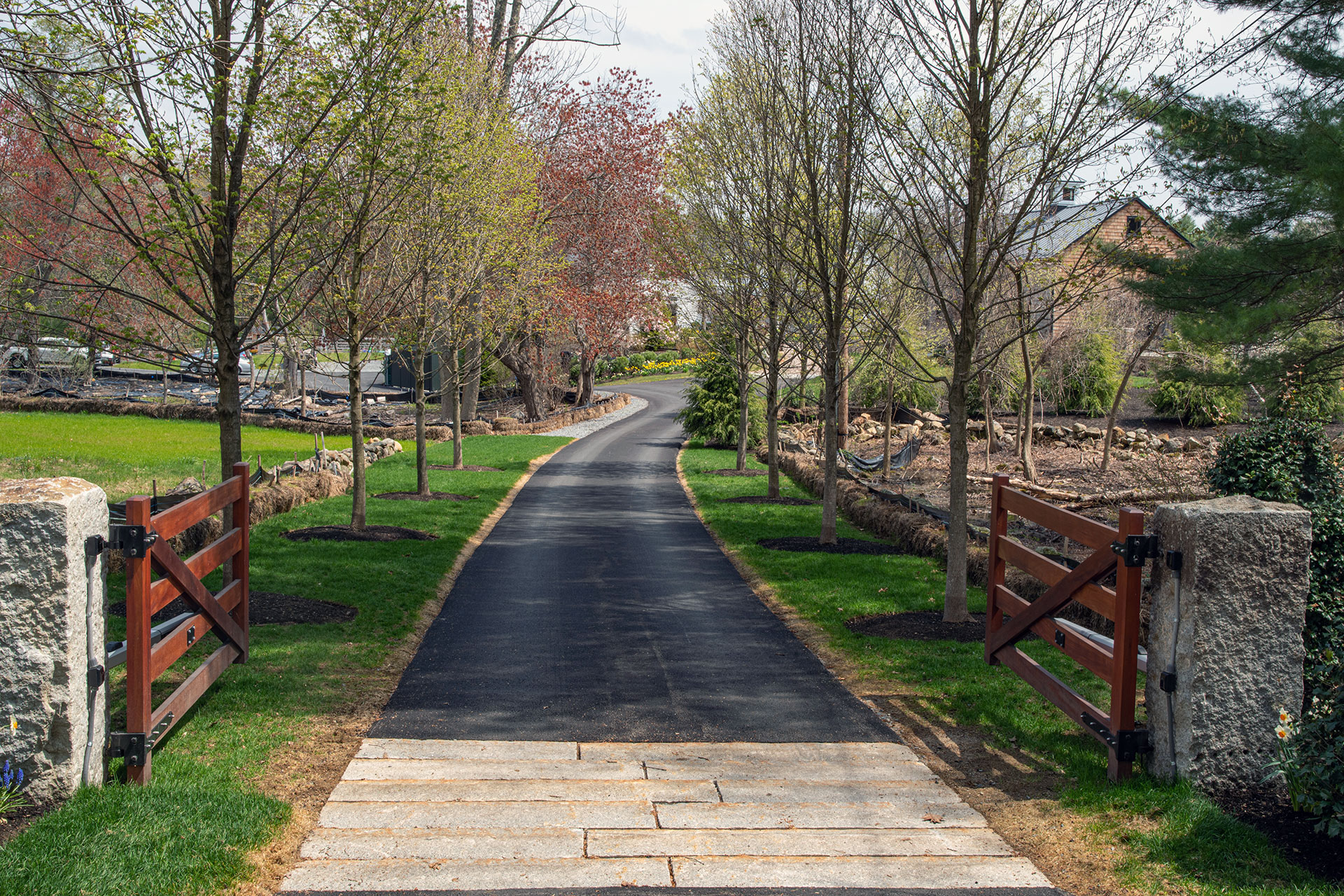 A smooth asphalt driveway is elevated by wood gates and granite posts, creating a tree-lined entry that balances everyday function with crafted details.