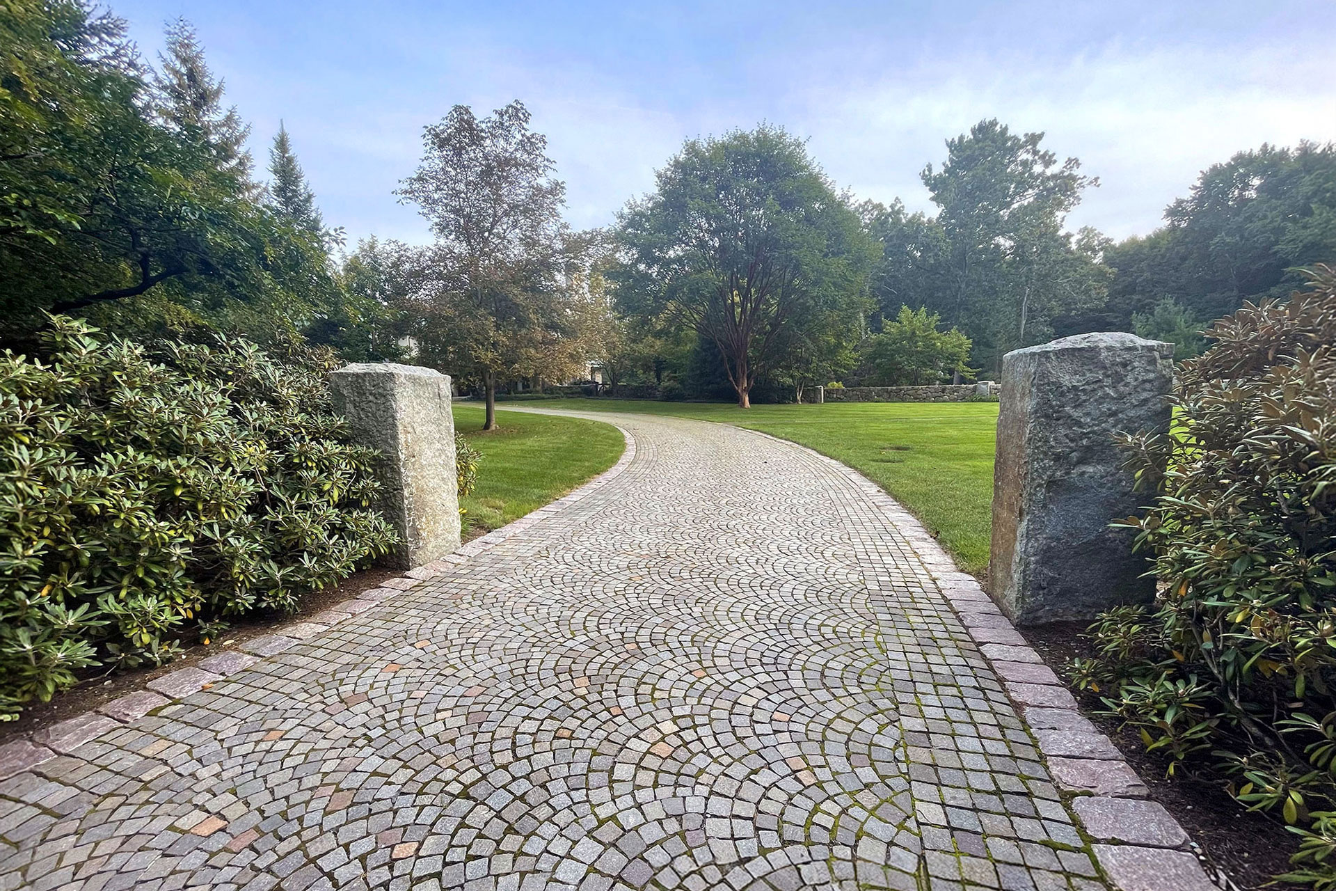 Curved cobblestone pavers lead to granite entry pillars, blending classic materials with a formal approach that balances visual weight, traction, and long-term durability.