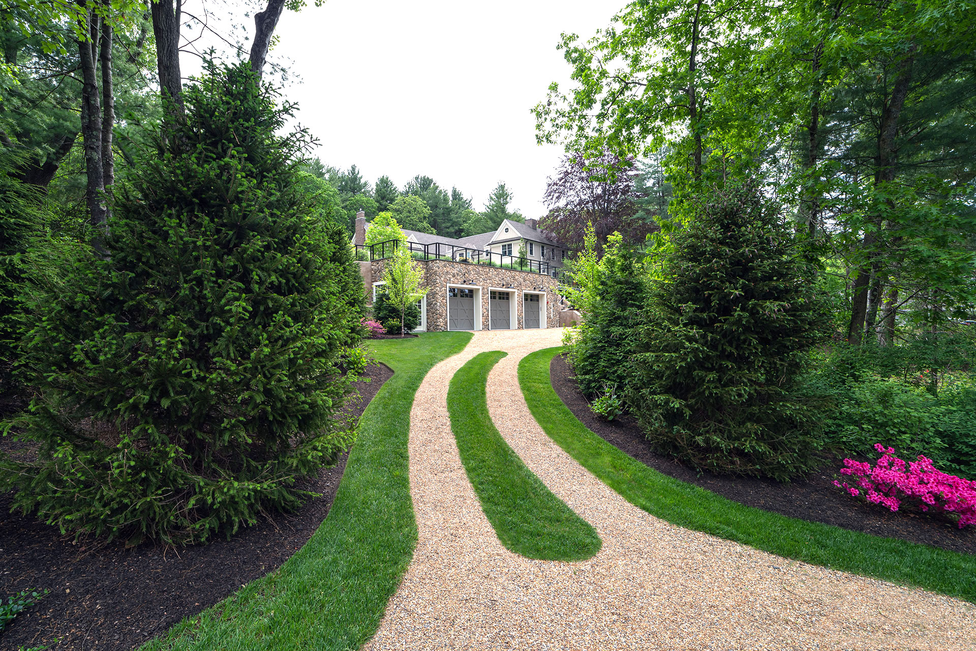 A double-track gravel driveway with a grass centerline reduces visual impact, guiding vehicles efficiently through a woodland setting toward a detached garage.
