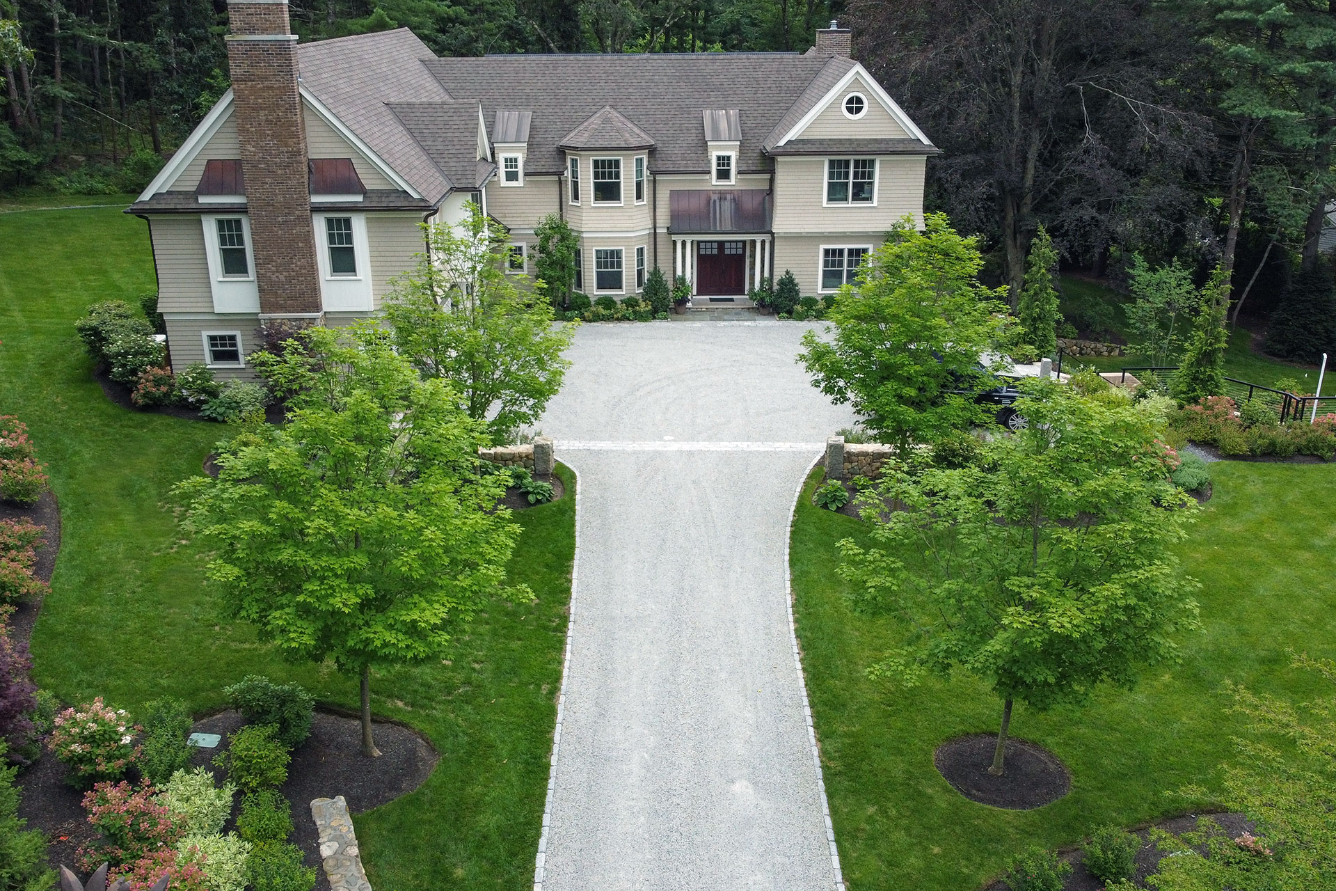 In Concord, MA, a symmetrical gravel driveway is framed by granite pillars, creating a formal, balanced approach that emphasizes proportion and long-term durability.