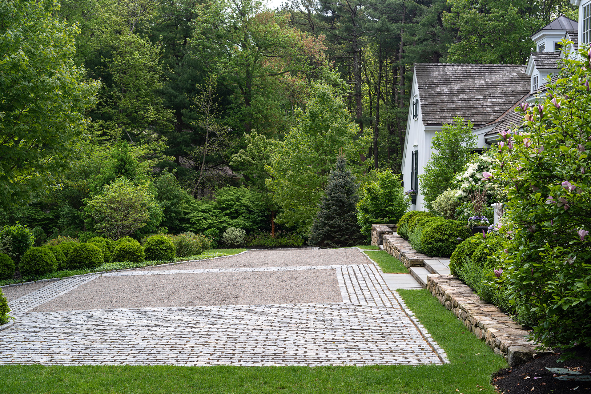 Gravel paving with cobblestone inlay leads toward stone steps, introducing subtle pattern and texture that elevates a formal entry without overwhelming it.