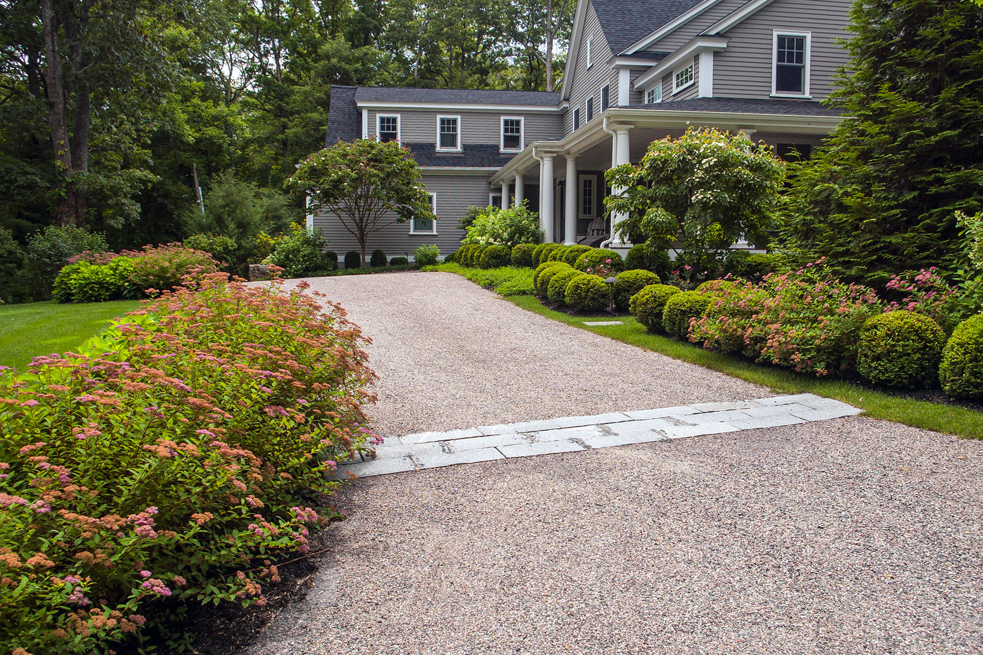 A gravel driveway bordered by layered perennials softens the approach to a colonial home, blending seasonal color with a practical, low-maintenance surface.
