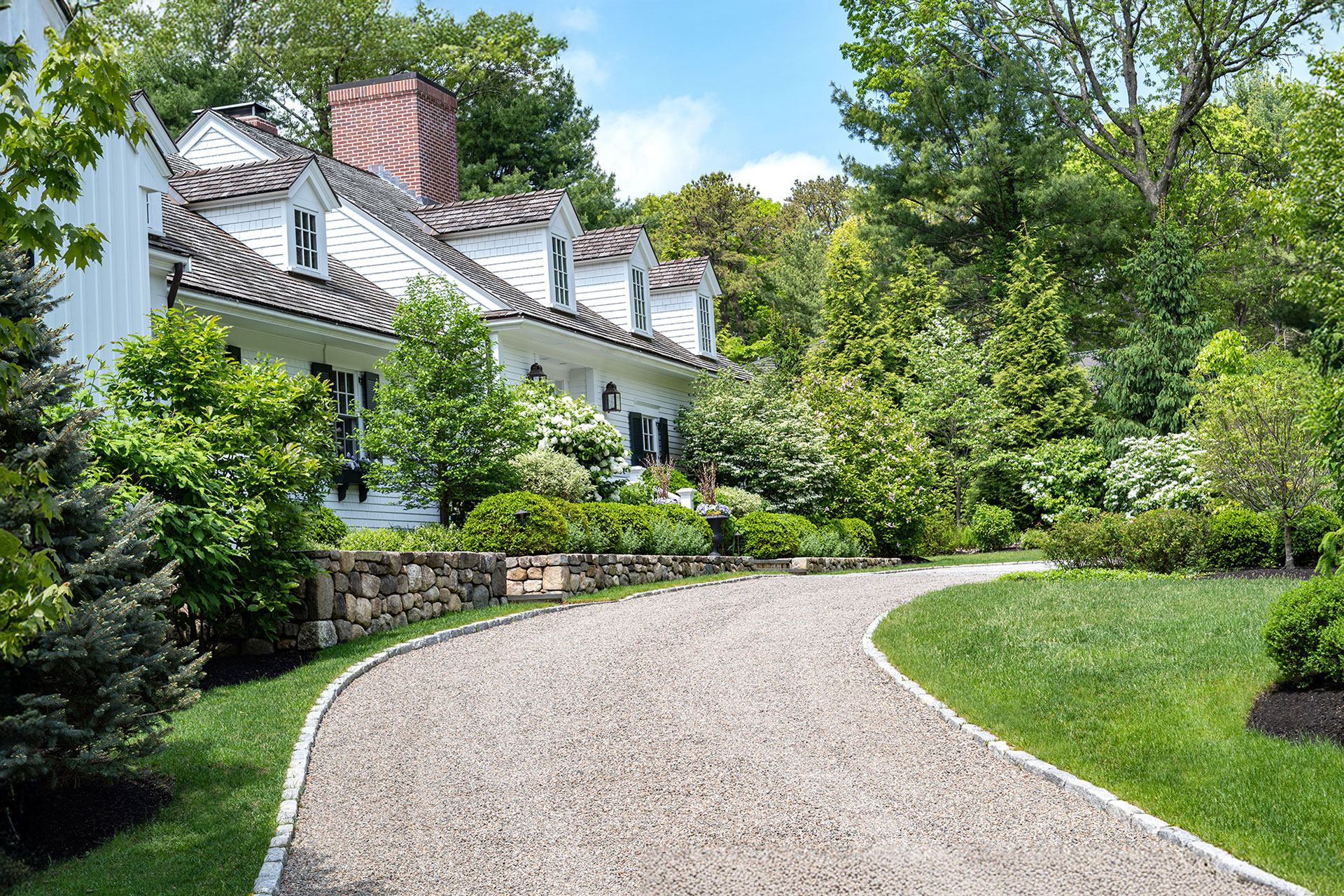 This tree-lined asphalt driveway pairs a smooth driving surface with wood gates and granite posts, creating a refined yet practical entry that feels intentional and welcoming.