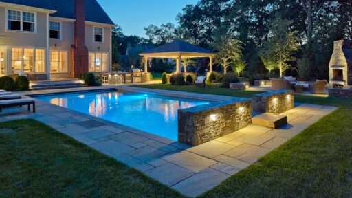 Landscape lighting adds warmth, depth, and definition, making the pool, stonework, and surrounding trees more inviting after dark.