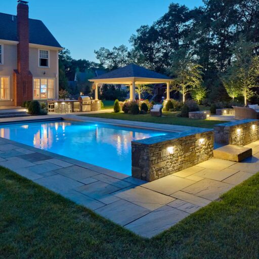 Landscape lighting adds warmth, depth, and definition, making the pool, stonework, and surrounding trees more inviting after dark.