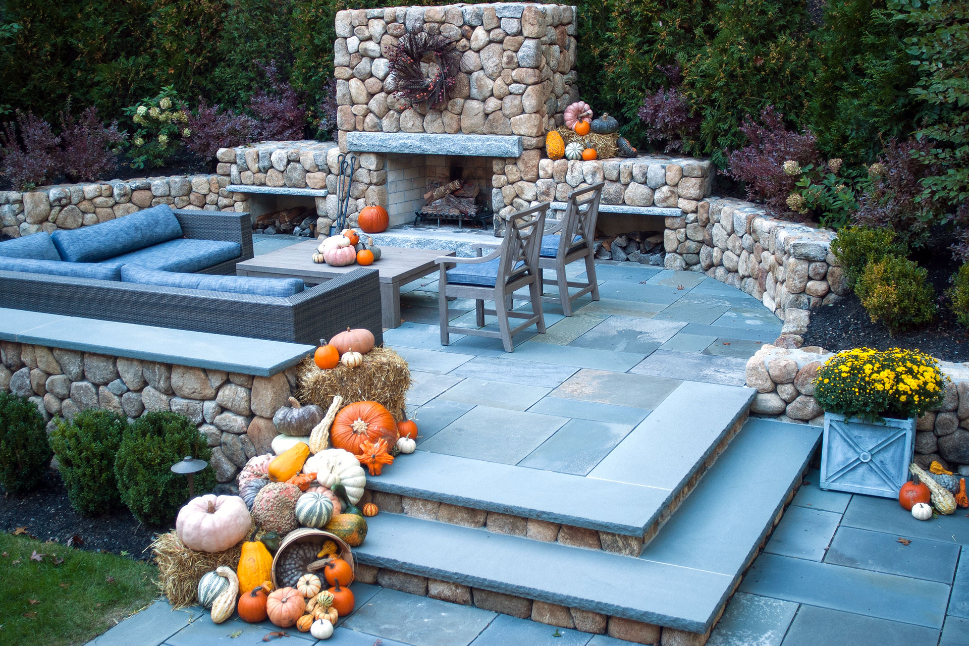 Pumpkins and hay bales surround a fieldstone fireplace on the patio, extending autumn style into outdoor living and entertaining spaces.