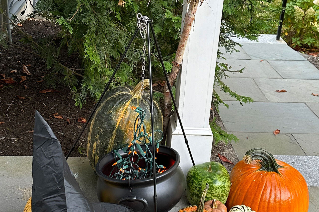 A cauldron planter paired with gourds and a witch hat adds a whimsical Halloween accent along the front walk, balancing spooky fun with landscape design details.