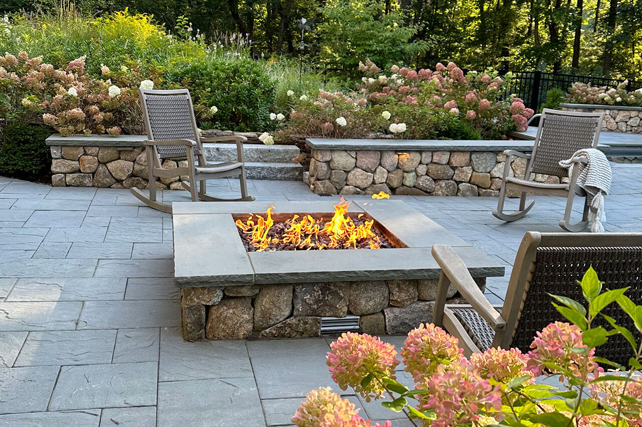 This square masonry fire pit in Sudbury, MA is framed by stone walls to create a defined outdoor room, offering durability, safety, and a comfortable focal point for year-round enjoyment.