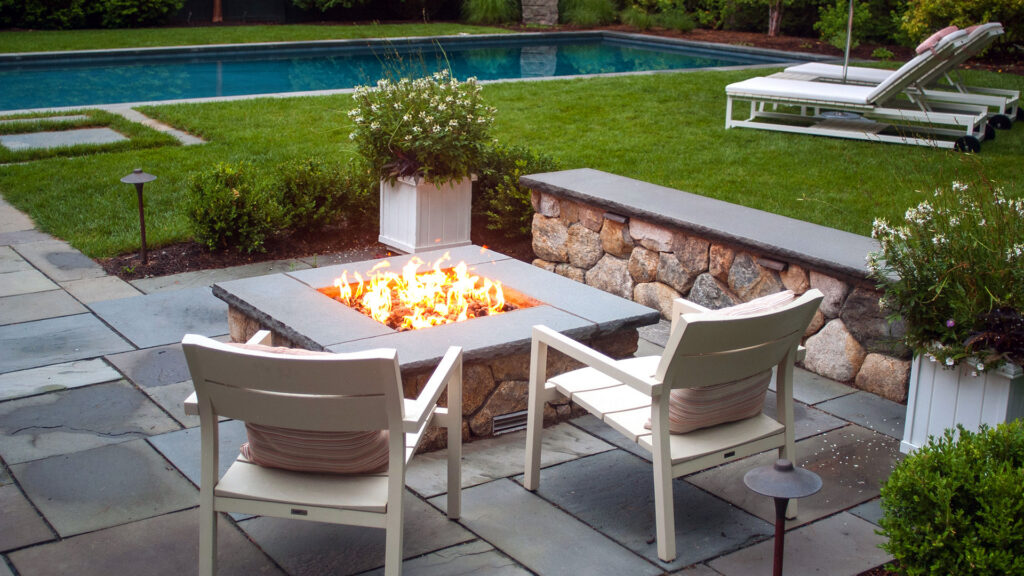 A square stone fire pit anchors this bluestone terrace beside the pool, offering clean lines, durable materials, and a central gathering point that enhances both day and evening use.