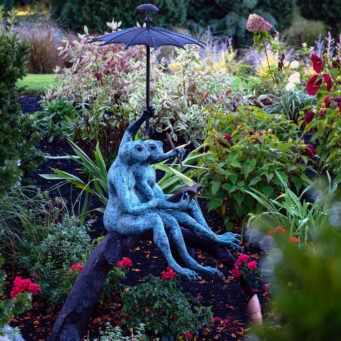 A bronze frog statue holding an umbrella brings humor and personality to the planting bed, its rich patina contrasting beautifully with fresh greens and flowers.