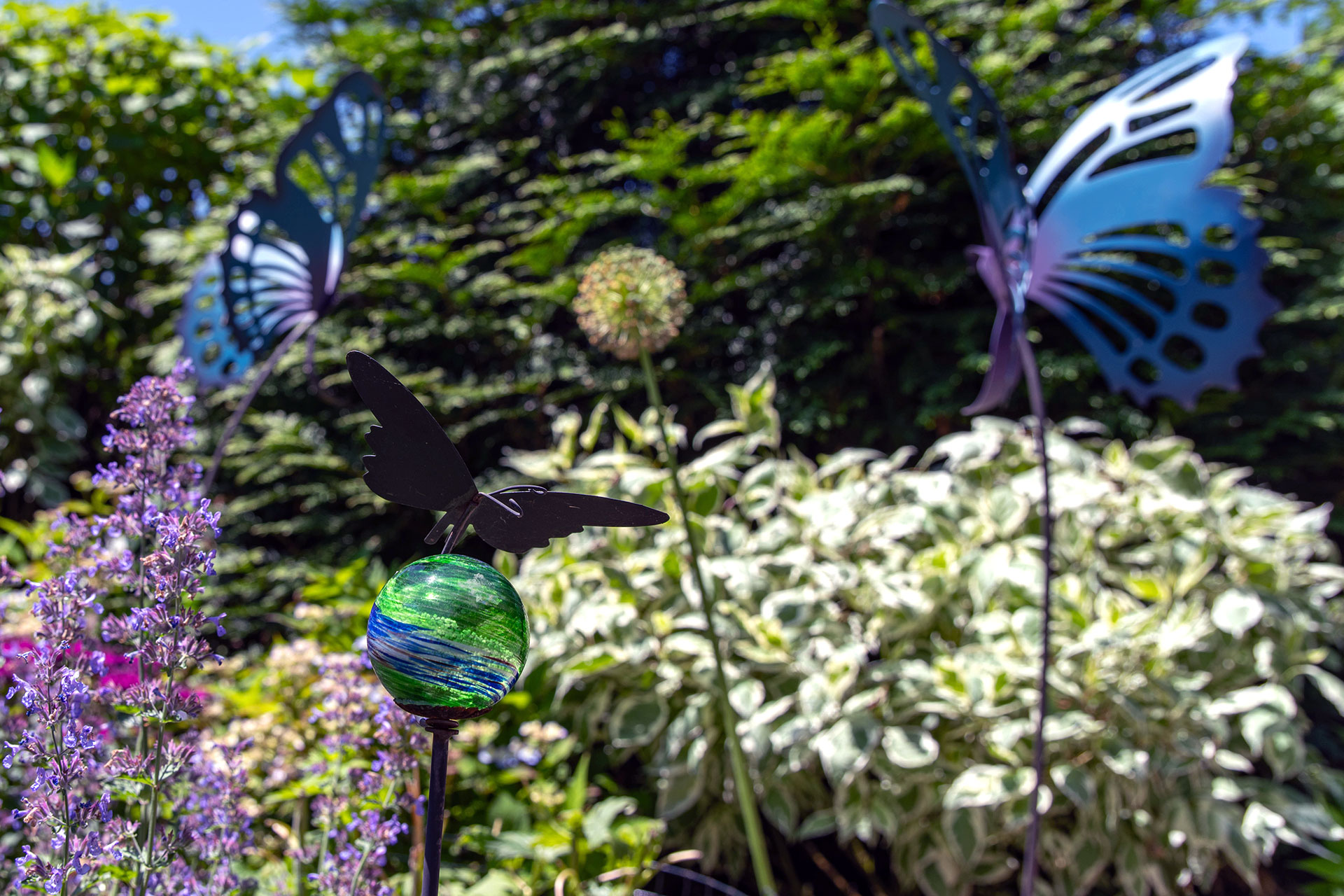 Colorful glass butterfly stakes flutter above perennial plantings, catching sunlight and adding vibrant blues and greens that animate the garden throughout the season.