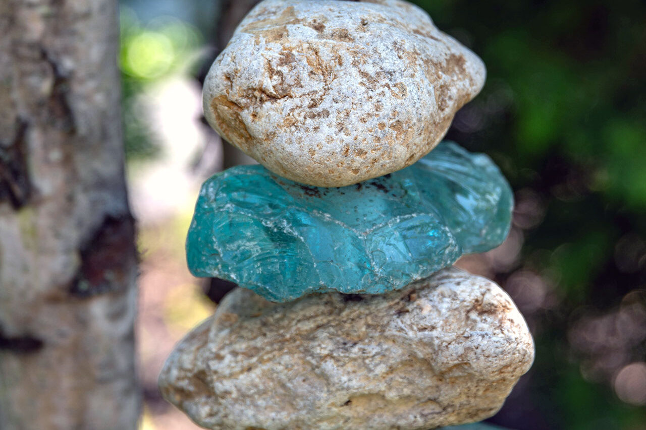 A stacked stone and glass cairn sculpture introduces vertical interest to the woodland garden, combining translucent blues with natural stone for a quiet, meditative focal point.